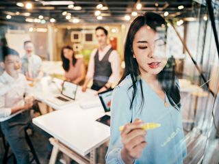 Asian woman writing on a glass wall in a room full of people