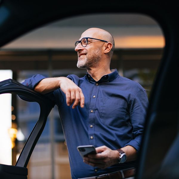 A man stands next to a car, casually leaning against the open door while holding a smartphone, showcasing a modern urban setting.