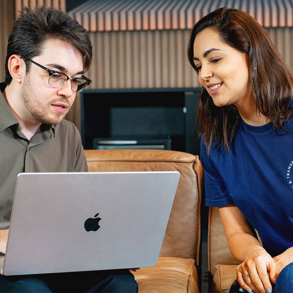A man and a woman in a casual office setting look at a laptop screen together while sitting on a brown leather couch.