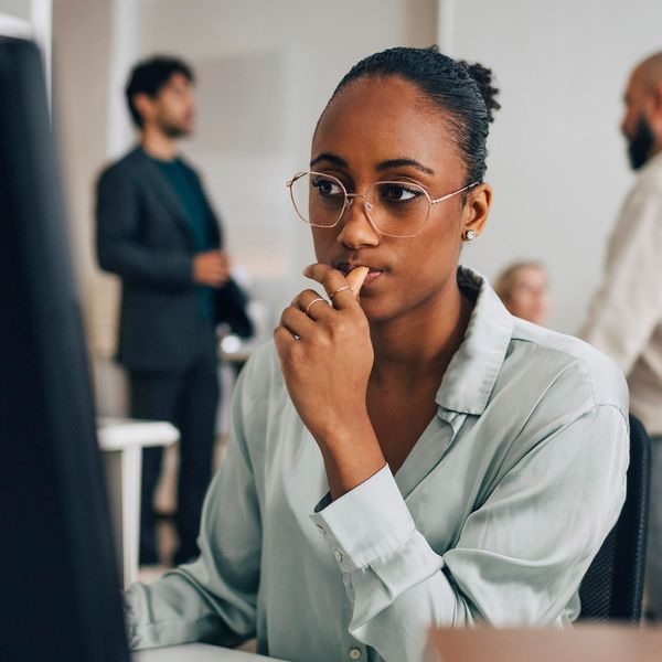 A young Black woman wearing glasses and a light green shirt looks thoughtfully at a computer screen in a bright office while colleagues converse in the background.