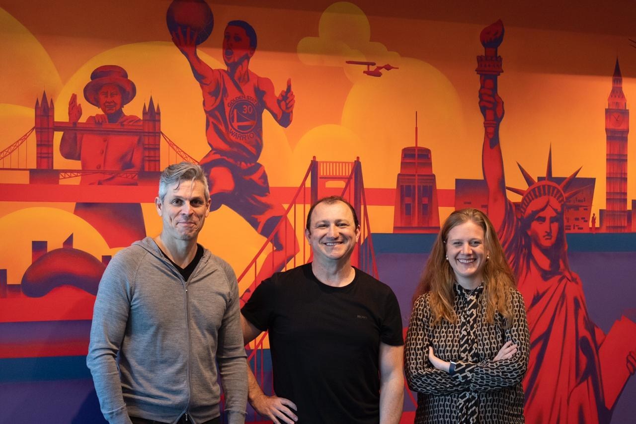 CI&T founders Bruno Guicardi and Cesar Gon with EMEA director Carolina Wosiack smiling and standing in front of a mural